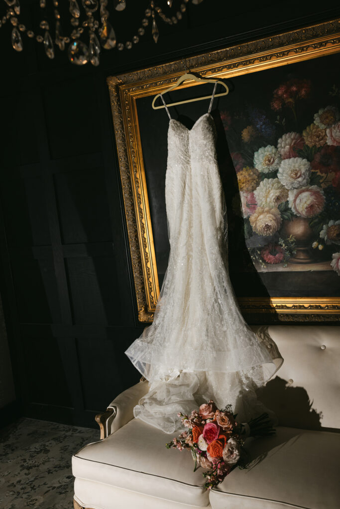 Bridal gown hanging in the suite at Edinburgh West in Greenville, SC, with sequin lace details, vintage floral painting, chandelier, and bridal bouquet on white sofa – Dani Lacey Photographs