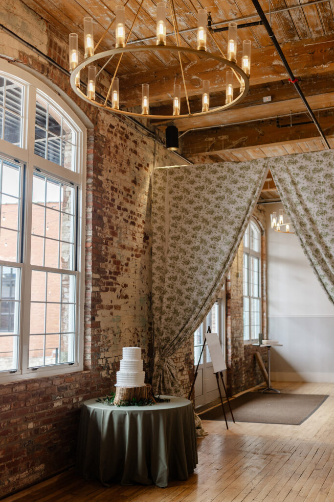 Wedding cake table with white tiered cake on wood slice inside rustic brick venue with chandelier and draped curtains in Greenville, SC – Dani Lacey Photographs