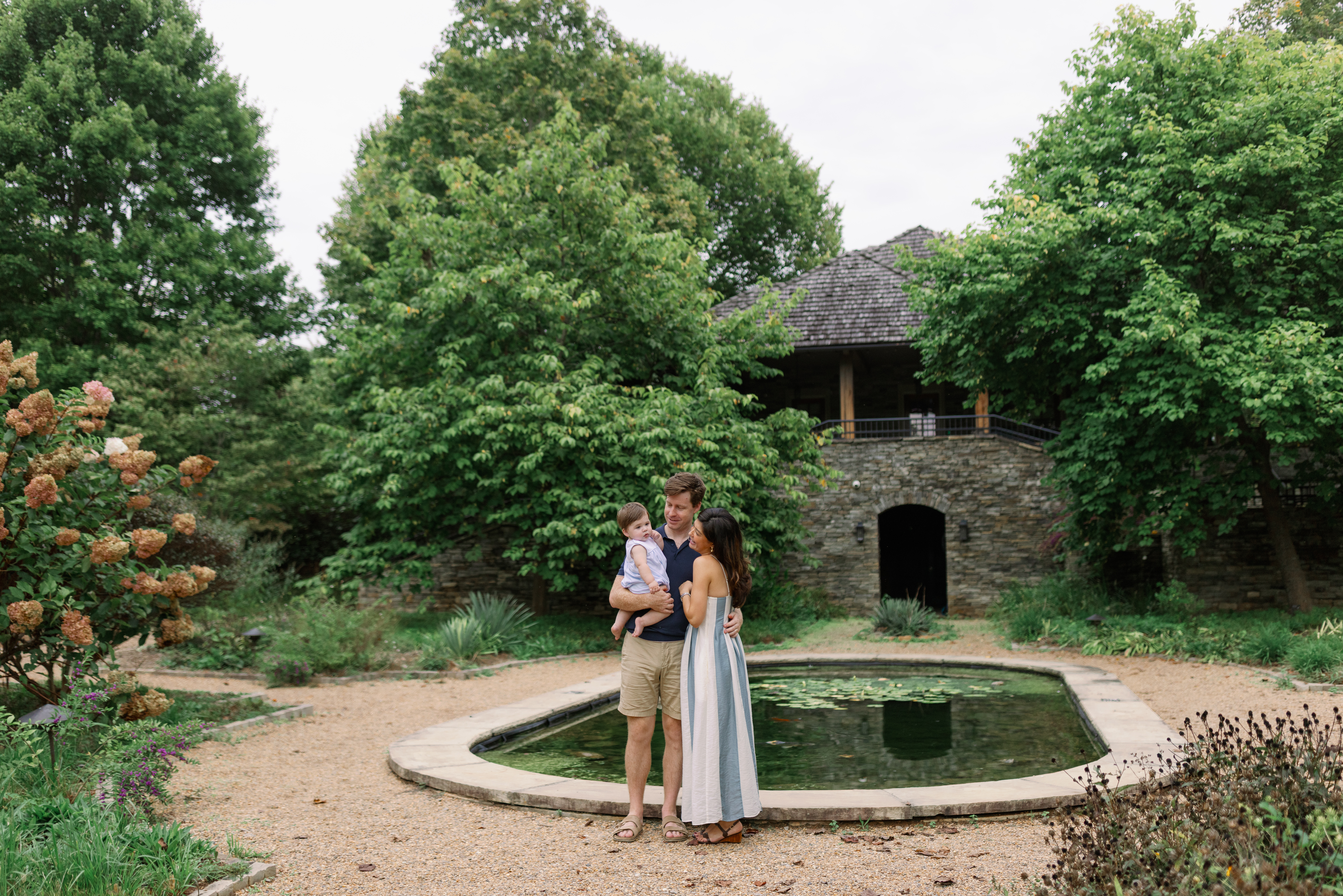 A family stands together by the pond with the stone house in the background during Greenville SC Family Portraits at Furman University. Dani Lacey Photographs.