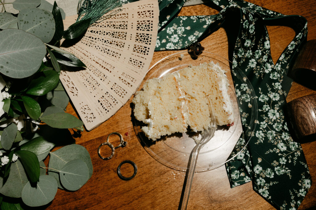 Flat lay of wedding rings, floral tie, wood fan, and a slice of wedding cake at Judson Mill wedding in Greenville SC – Dani Lacey Photographs