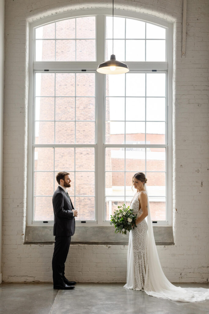 Bride and groom facing each other during first look in front of large industrial windows at Judson Mill Greenville.