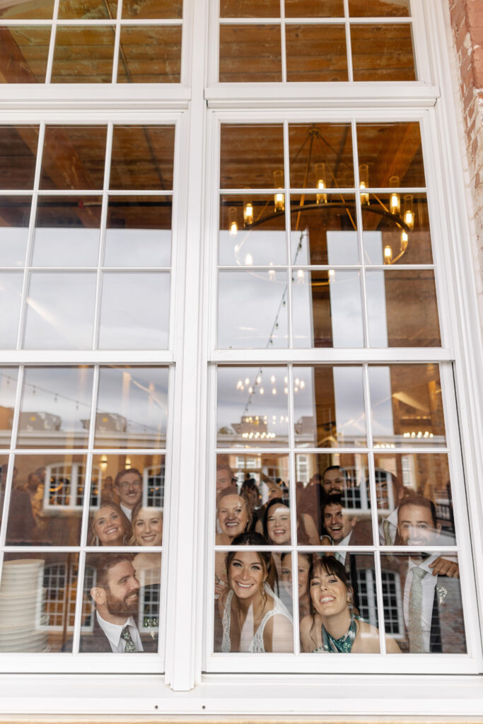 Bridal party peers out of a large industrial window at Judson Mill, creating a playful and unique group wedding portrait. Dani Lacey Photographs.