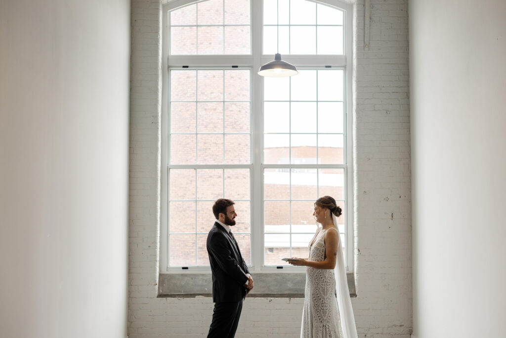 Bride and groom stand together during their first look by the windows of Judson Mill Greenville.