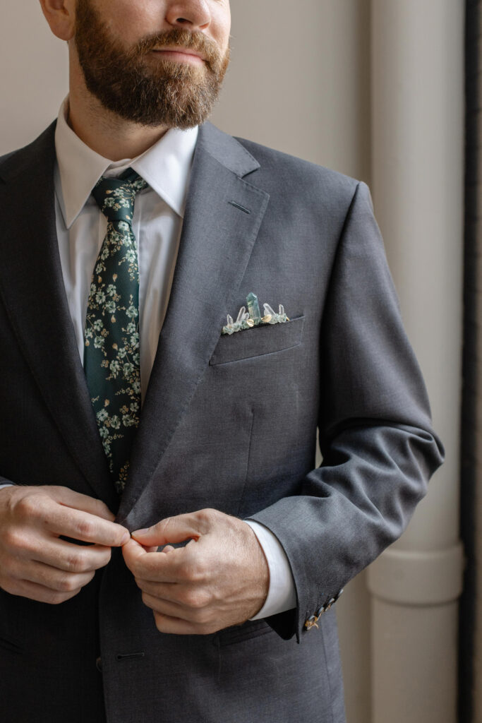 Close-up of groom adjusting his suit jacket with floral tie and crystal pocket square at Judson Mill wedding captured by Greenville wedding photographer Dani Lacey.
