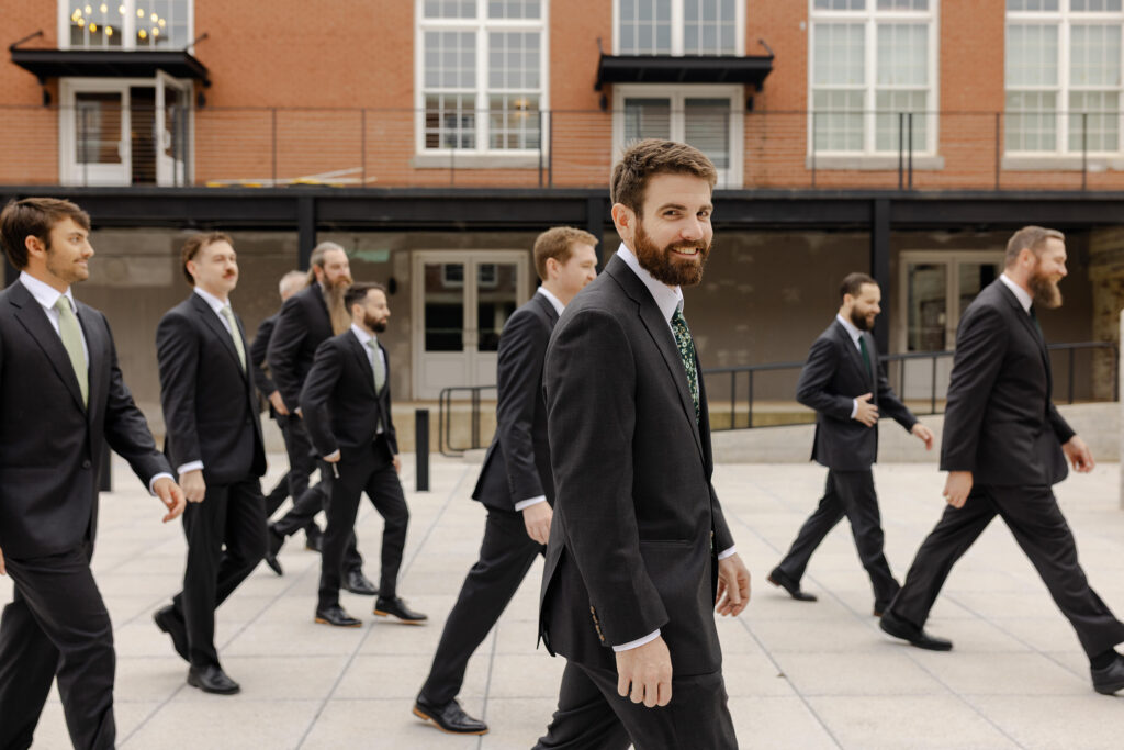 Groom smiling at the camera while walking with his groomsmen outside Judson Mill in Greenville SC.