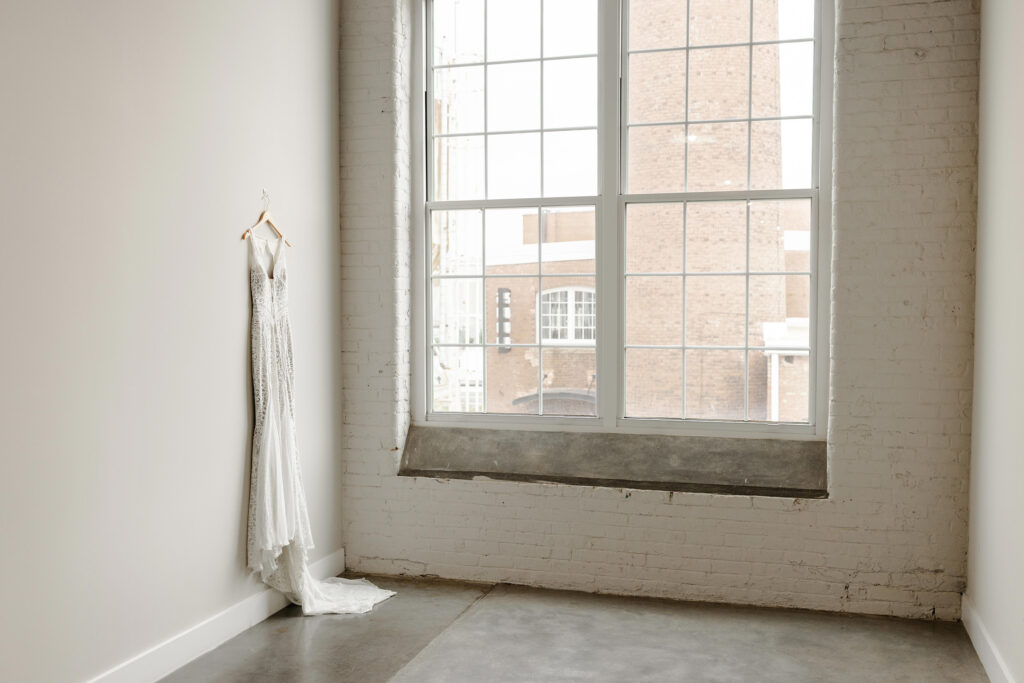 A lace wedding gown hanging against a white brick wall inside Judson Mill in Greenville SC, captured by Dani Lacey Photographs.