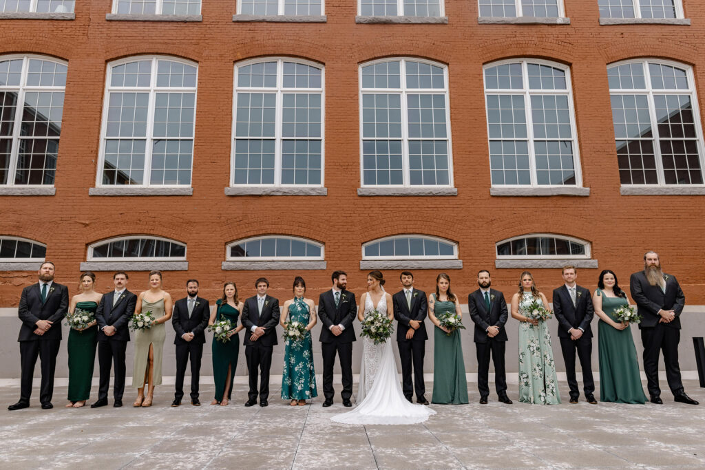Full wedding party lined up against the industrial brick walls of Judson Mill in Greenville, SC, with the bride and groom at the center. Dani Lacey Photographs.