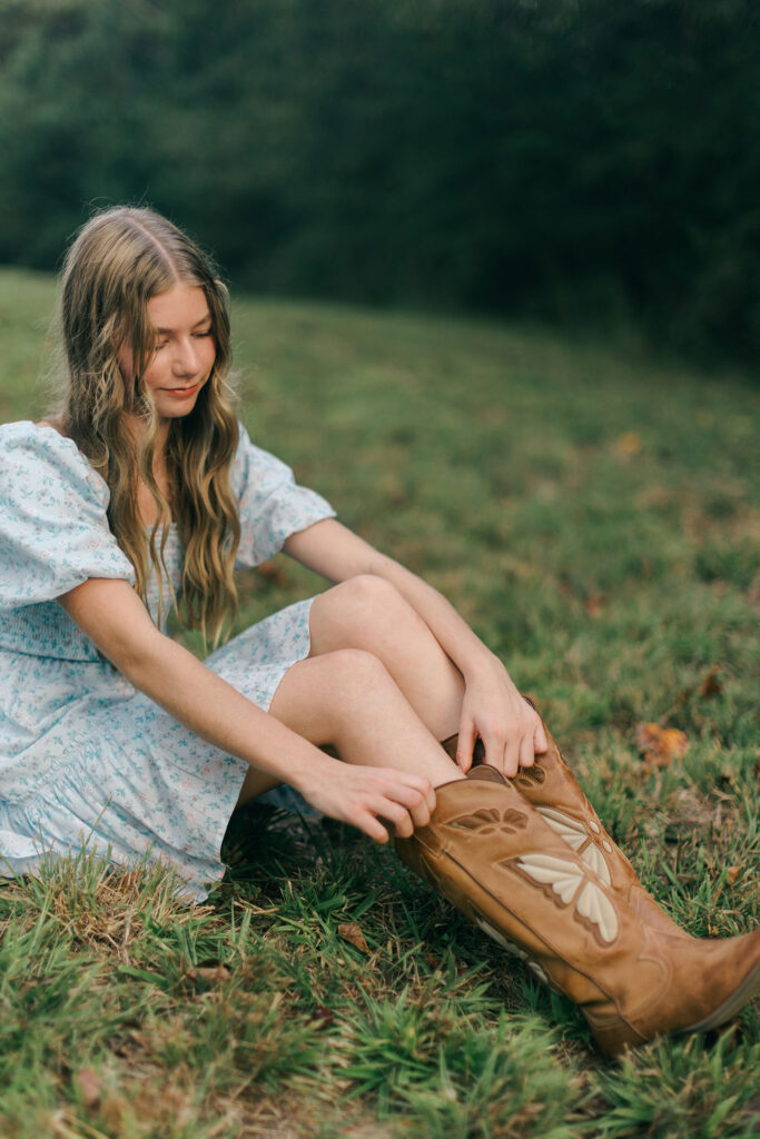Teen girl sitting in the grass adjusting her cowboy boots during an at-home family photo session in Greenville SC. Dani Lacey Photographs.