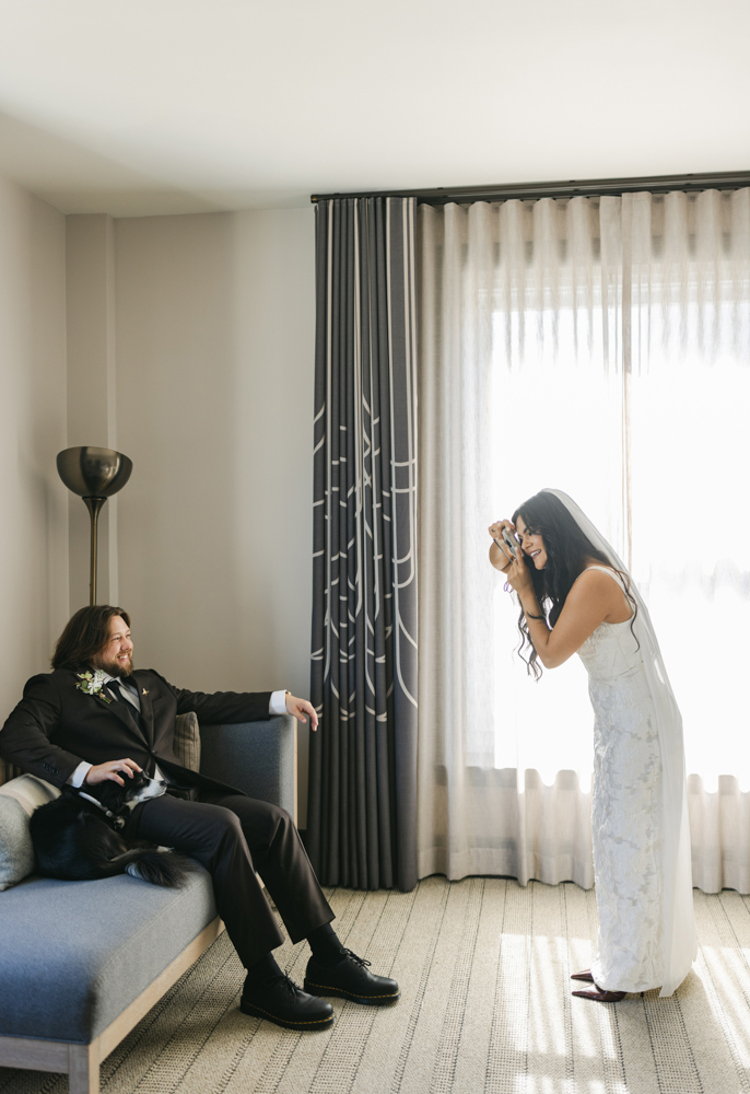 bride taking photo of groom in hotel room