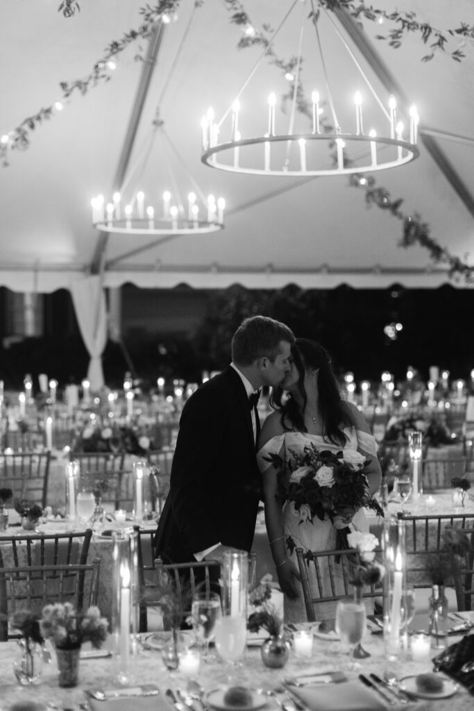 Bride and groom share a kiss beneath chandeliers inside an elegant candlelit wedding reception tent.