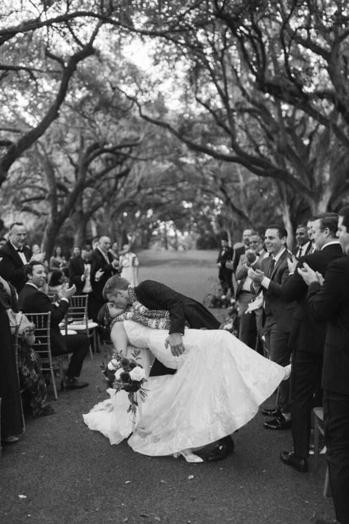 Groom dips and kisses his bride during an outdoor wedding ceremony as guests applaud.
