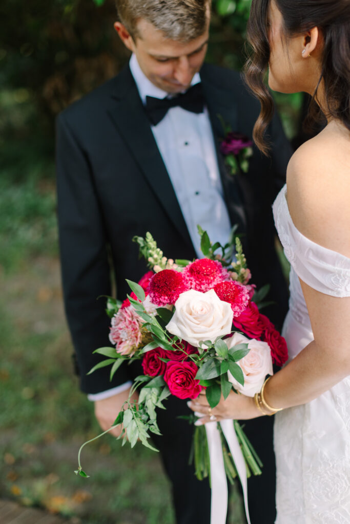 Bride holding a colorful bouquet of red, pink, and ivory flowers beside the groom in a black tuxedo.
