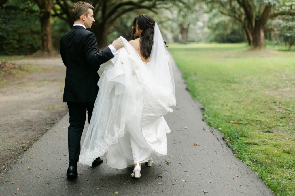 Groom carries his bride's wedding dress train as they walk together along a tree-lined path.