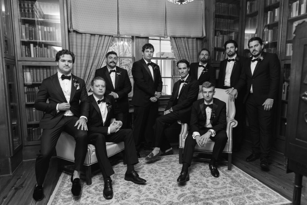 Eight groomsmen in black tuxedos pose for an editorial black and white wedding portrait inside a library.