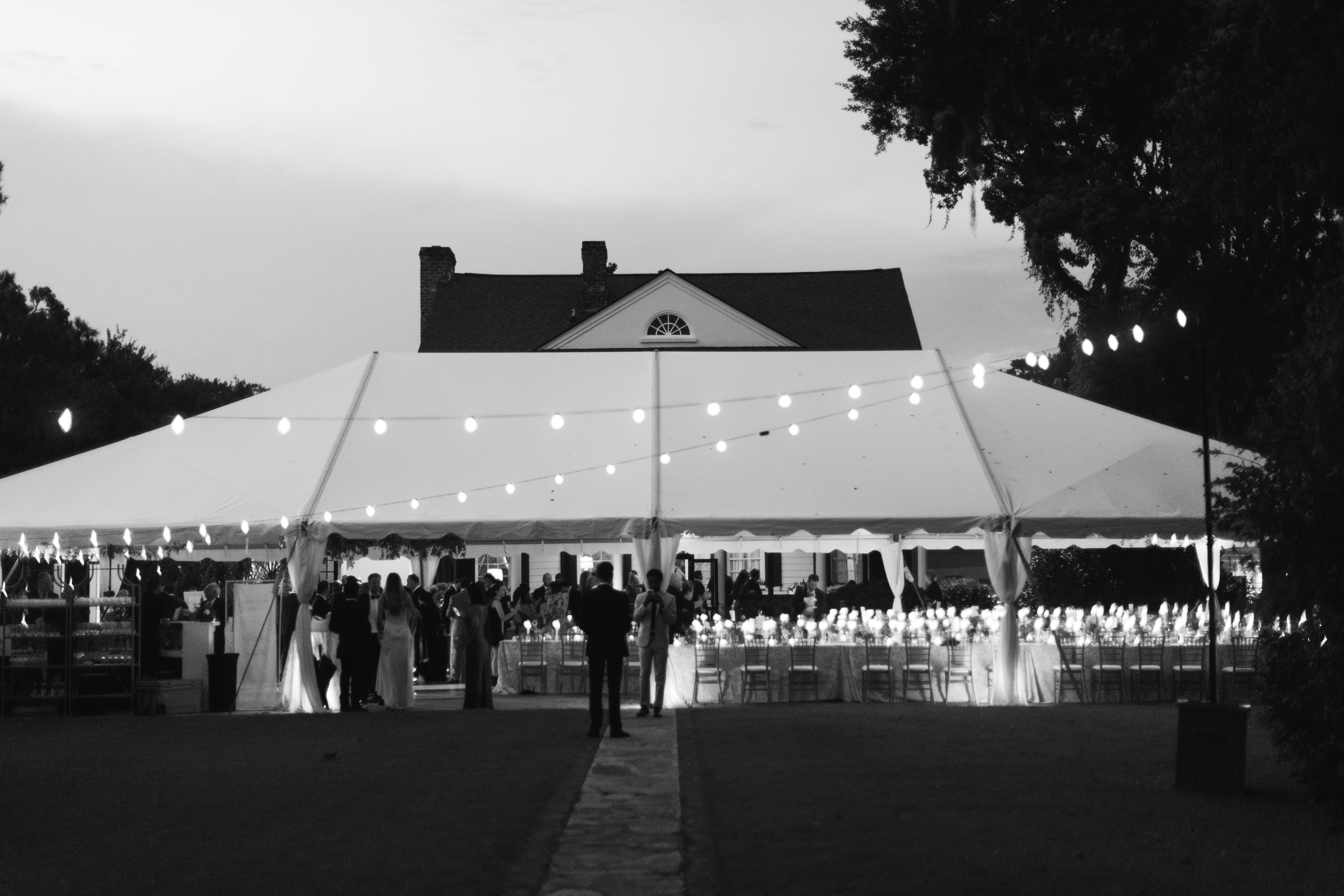 Large white wedding reception tent illuminated by string lights at dusk with guests gathering inside.
