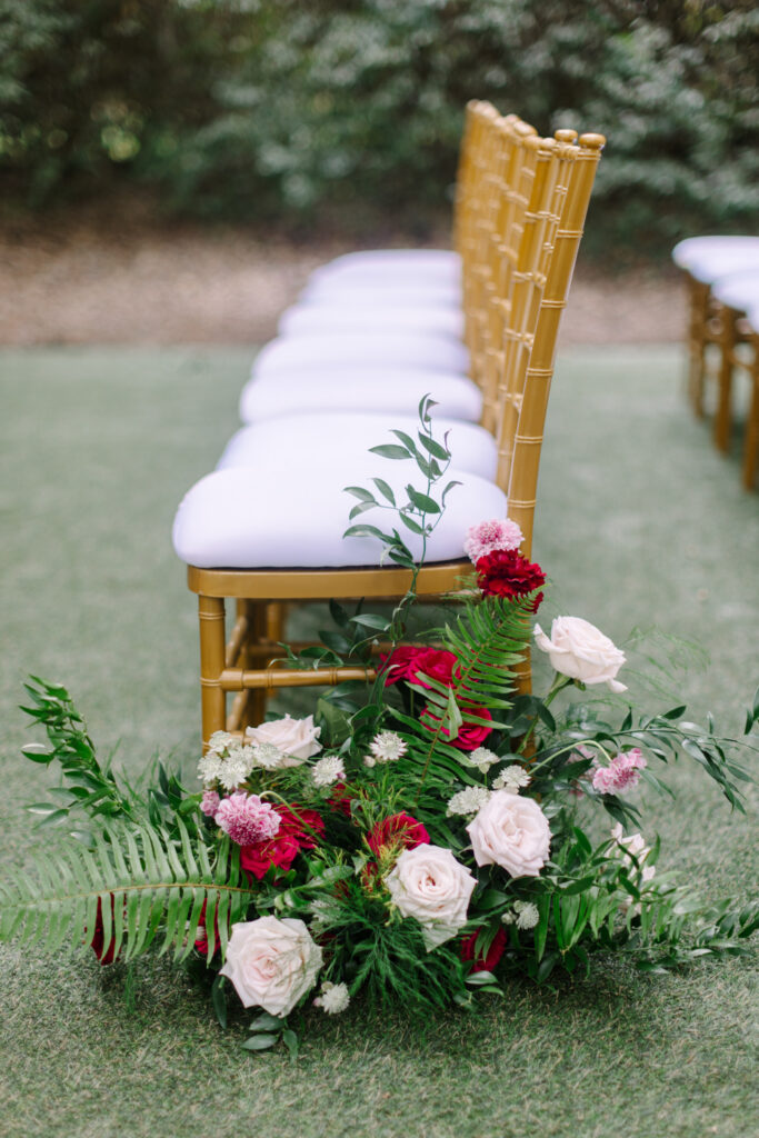 Red, blush, and ivory wedding flowers arranged beside gold ceremony chairs at an outdoor wedding.