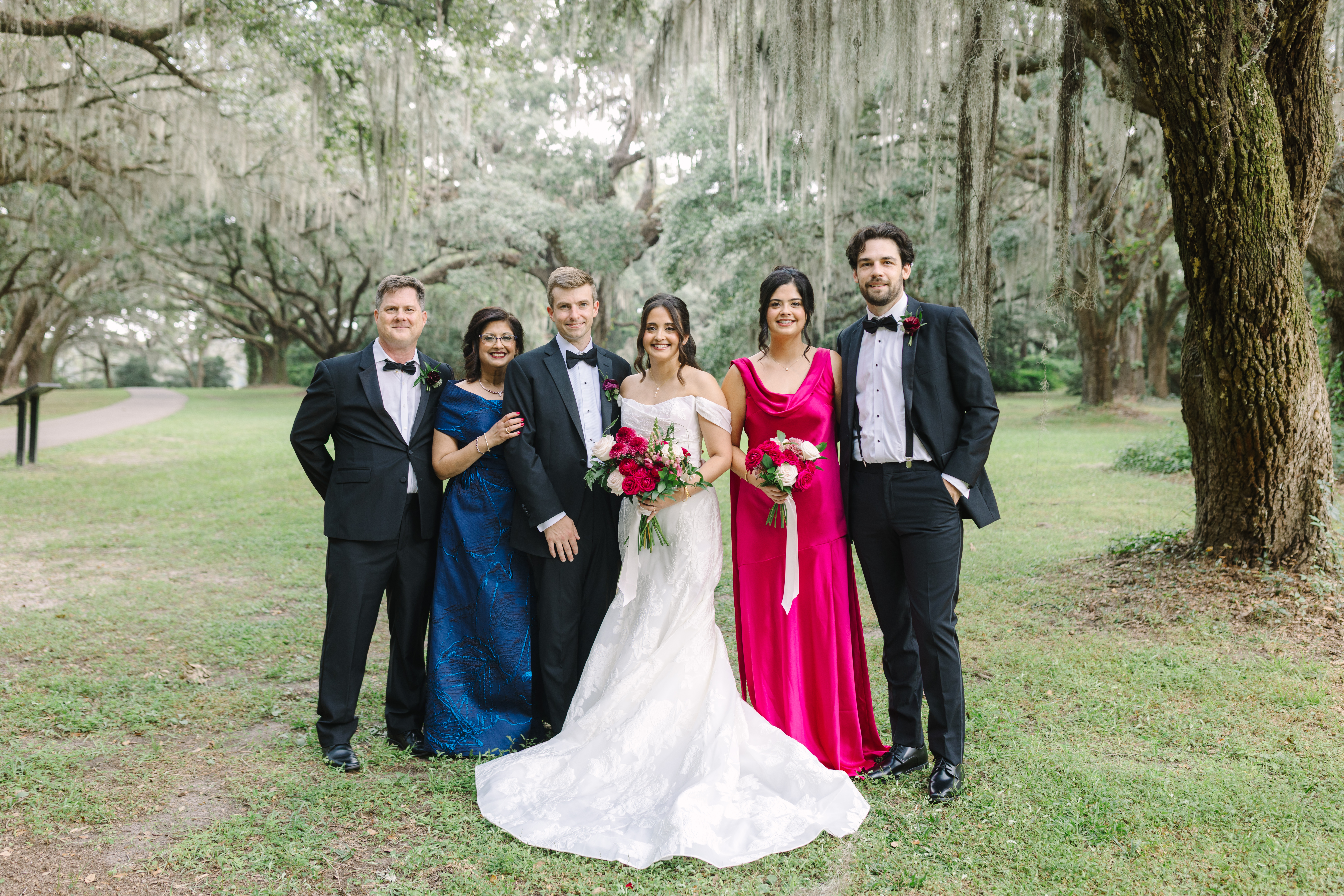 Bride and groom pose with immediate family members beneath Spanish moss-covered oak trees during an outdoor wedding.