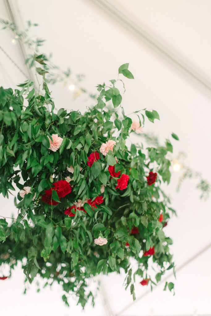 Large overhead wedding floral installation featuring lush greenery and red and pink flowers inside a white reception tent.